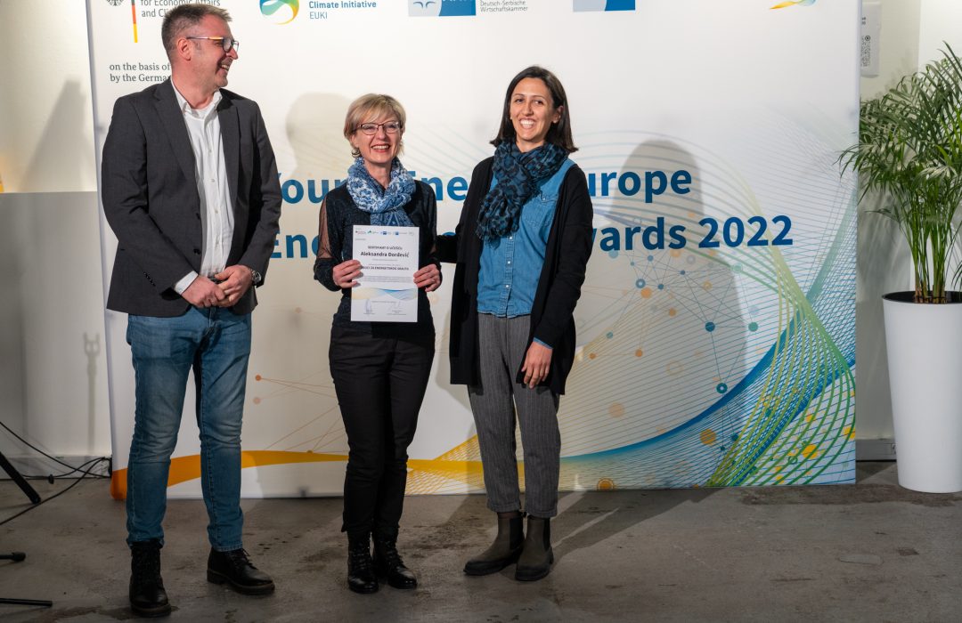 A man and two women standing in front of a Young Energy Europe roll-up, the woman in the middle is holding a certificate
