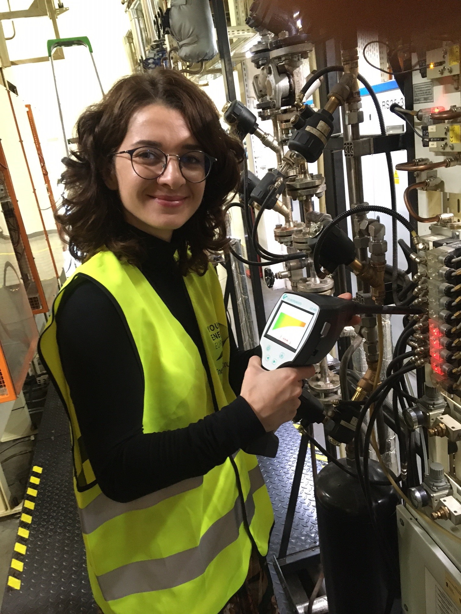 A woman in a yellow Energy Scout vest holding a leak detector to pipes, smiling at the camera