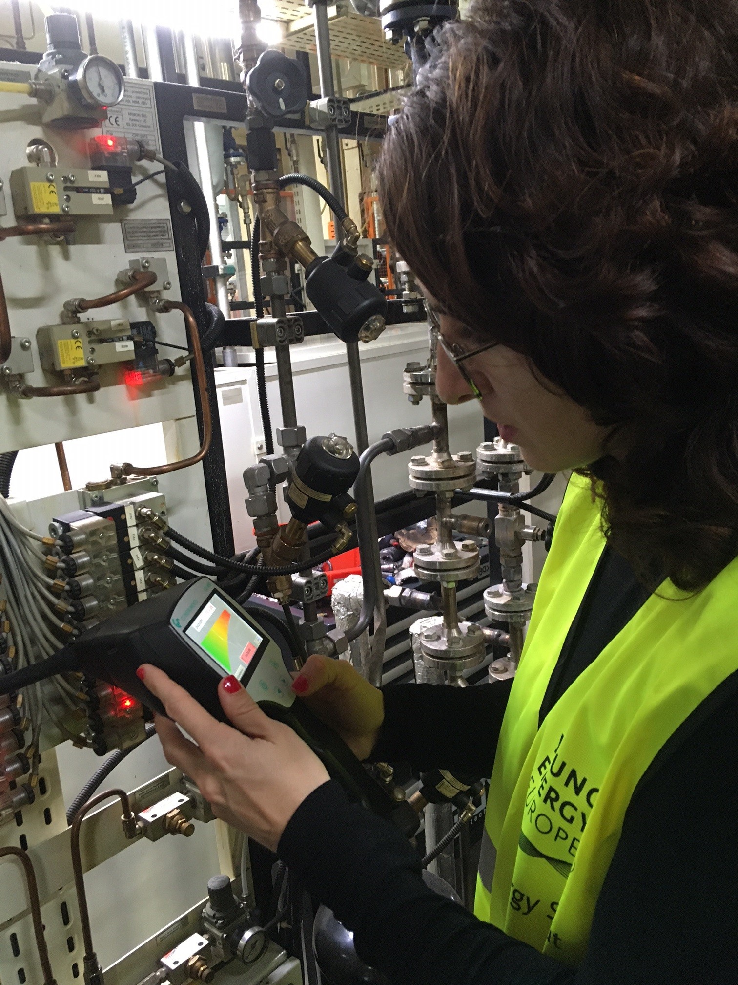 A woman in a yellow Energy Scout vest holding a leak detector to pipes, looking at the screen