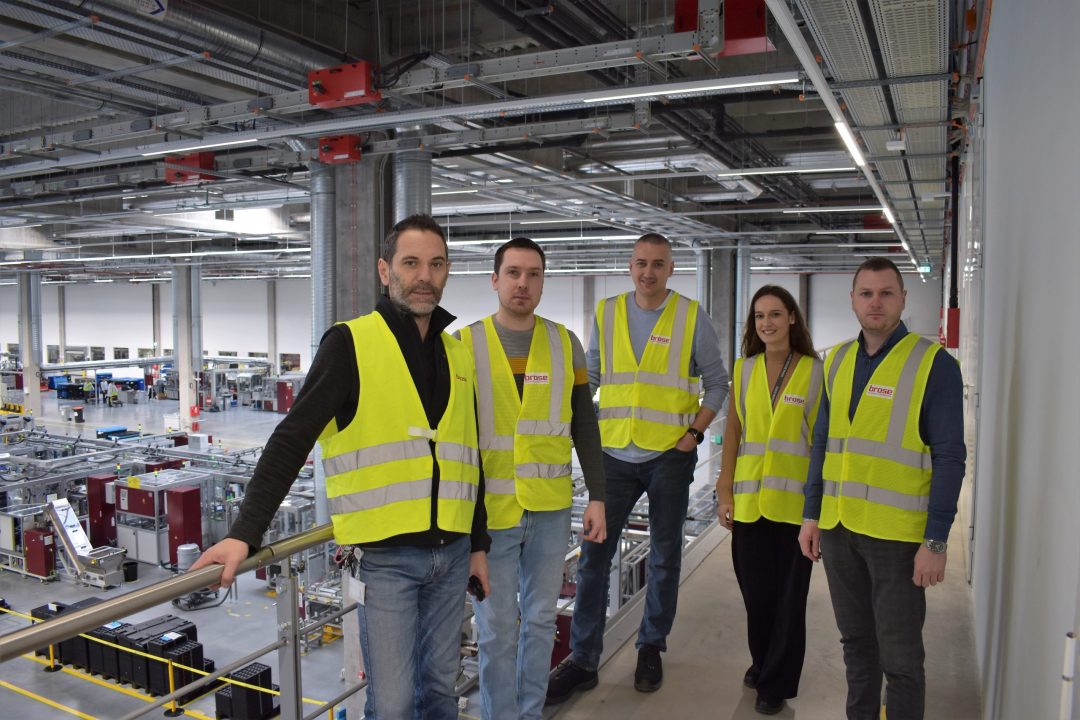 Eine Gruppe aus vier Männern und einer Frau stehen in in gelben Warnwesten nebeneinander und stehen in einer Produktionshalle