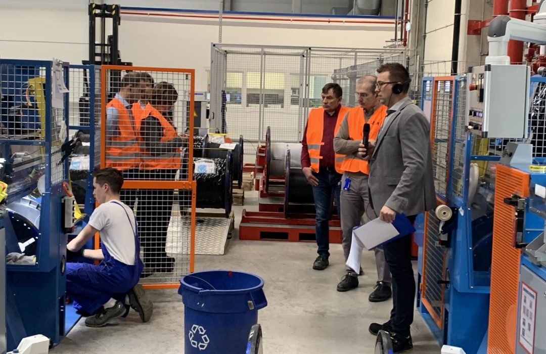 Five men in orange safety vests are in a production plant. One man with a microphone and headphones is explaining. Another man is operating a production machine.