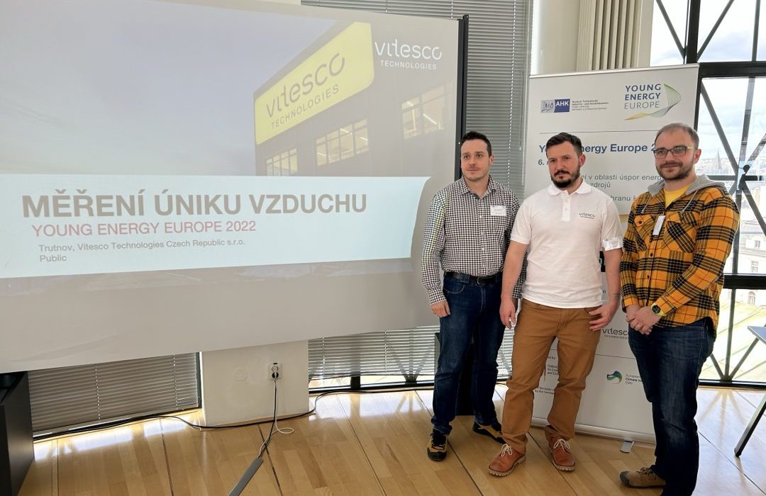 Three young men are standing on the right side of the picture in front of a screen with a PowerPoint presentation in Czech.
