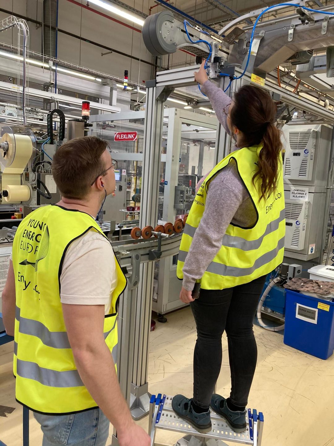 A man and a woman in yellow Young Energy Europe vests are next to the assembly line of a production room. The woman is pointing at the top of a connection from the assembly line with cables and gears, the man is also looking at it.