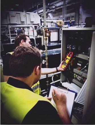 Two men in yellow Vests use an electricity measuring tool inside a machine box