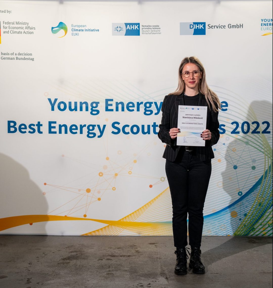 Eine junge Frau steht vor einer Wand auf der Young Energy Europe, EUKI, DIHK Service GmbH und AHK Serbien Logos abgebildet sind und hält eine Urkunde in der Hand.