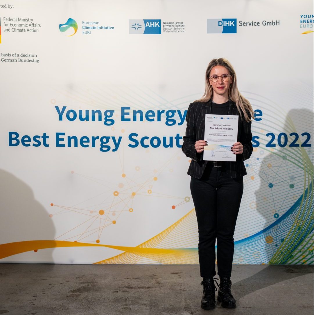 A young woman stands in front of a wall on which Young Energy Europe, EUKI, DIHK Service GmbH and AHK Serbia logos are displayed and holds a certificate in her hand.