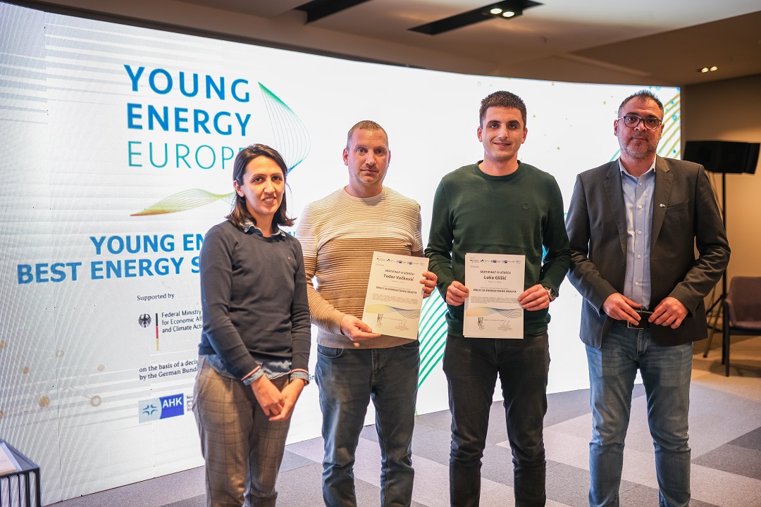 A woman and three men stand in front of a large display with Young Energy Europe branding, two of them holding up certificates
