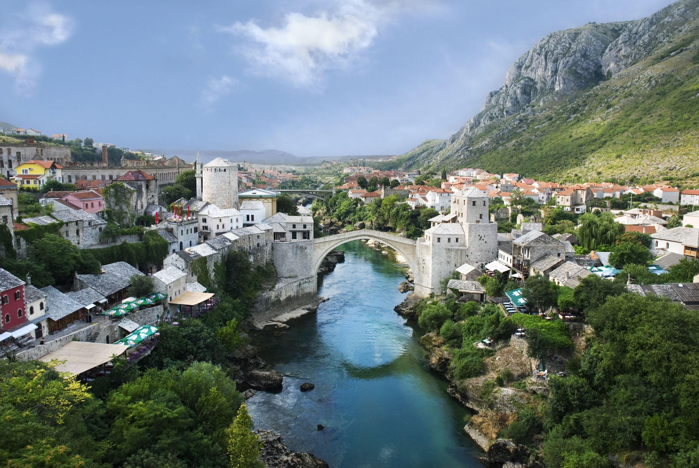 View of Mostar, Bosnia and Herzegovina, with the Stari Most bridge over the Neretva River, surrounded by stone buildings, greenery, and mountains