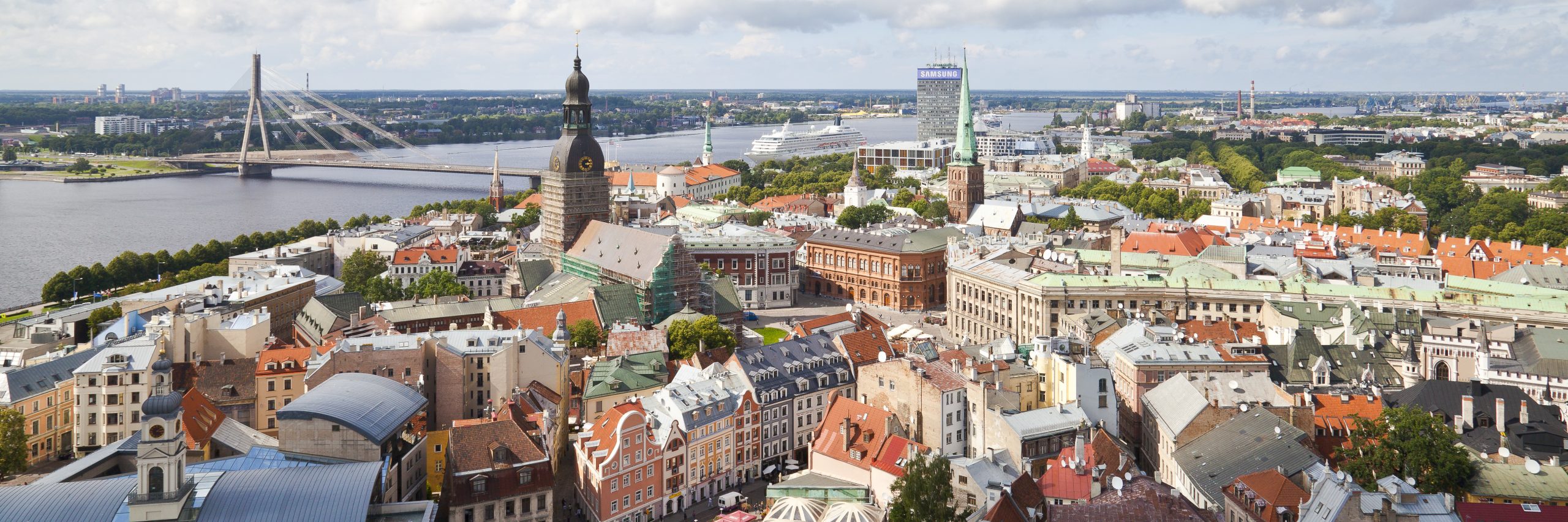 Aerial view of a city with a river and a prominent bridge. A large church with a spire and clock tower stands in the center. Modern buildings and greenery line the riverbanks under a partly cloudy sky