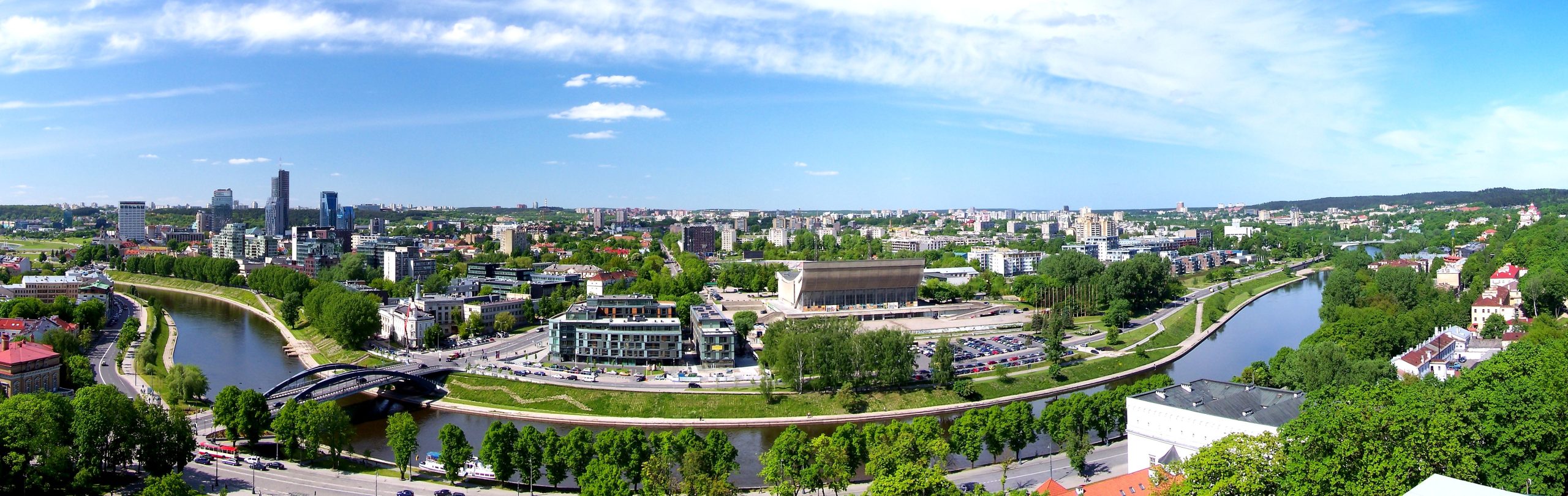 Panoramaansicht einer Stadt mit Fluss, Brücke und verschiedenen Gebäuden. Ein großes Gebäude mit Parkplatz steht zentral. Viel Grün und teils bewölkter Himmel