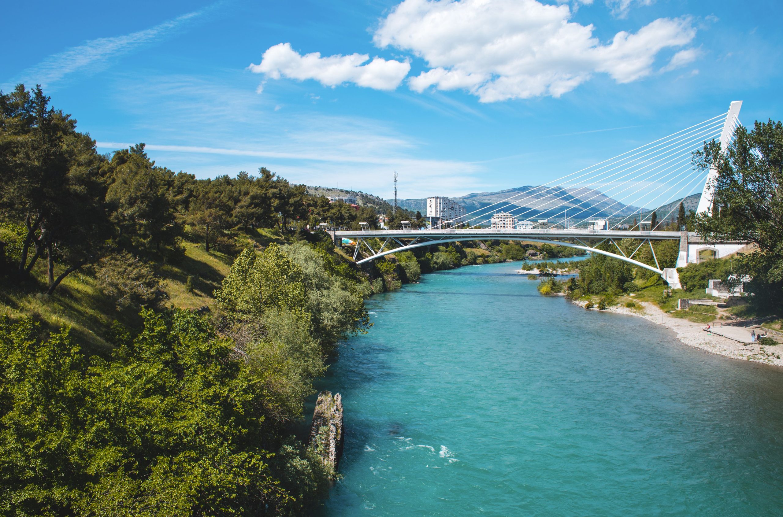 A cable-stayed bridge spans a river in a green landscape with trees, buildings, and mountains under a partly cloudy sky