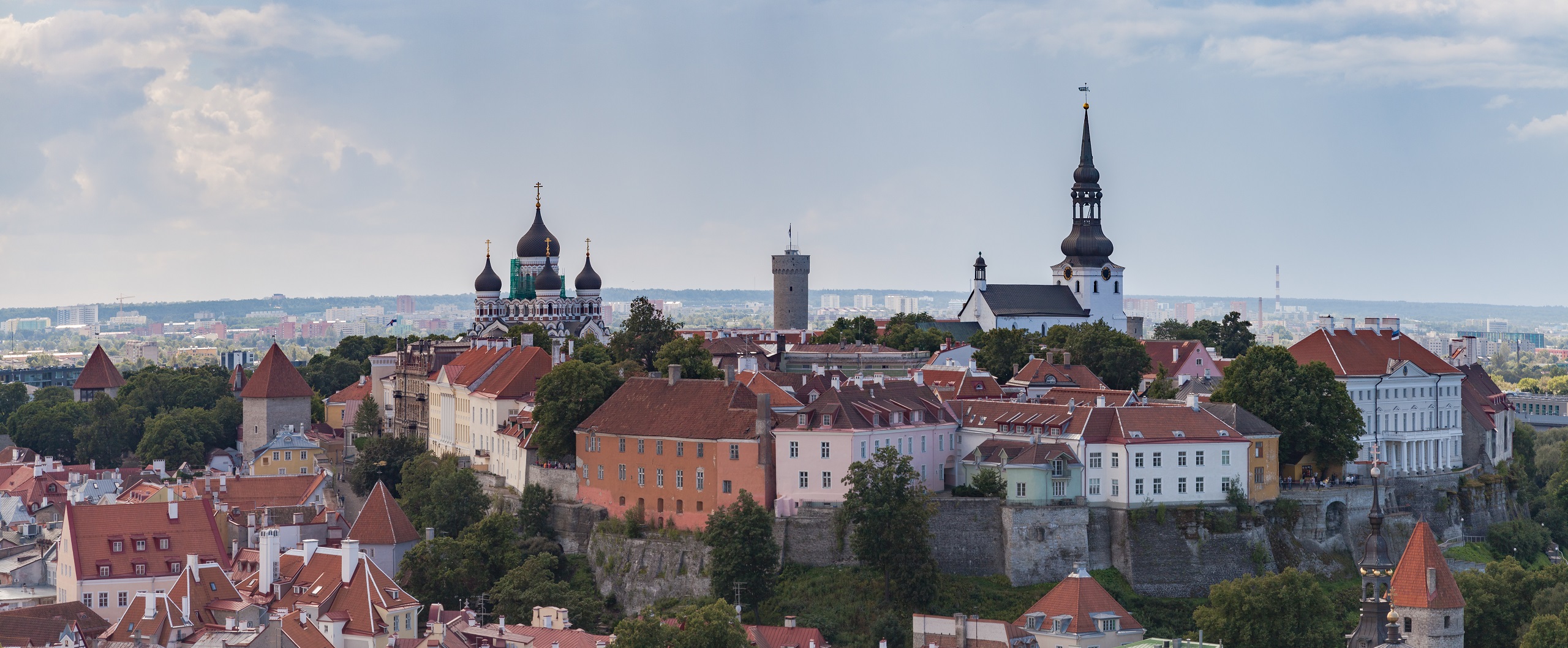 Panoramic view of a historic city with churches featuring onion domes and spires, colorful buildings with red roofs, and greenery surrounding the area