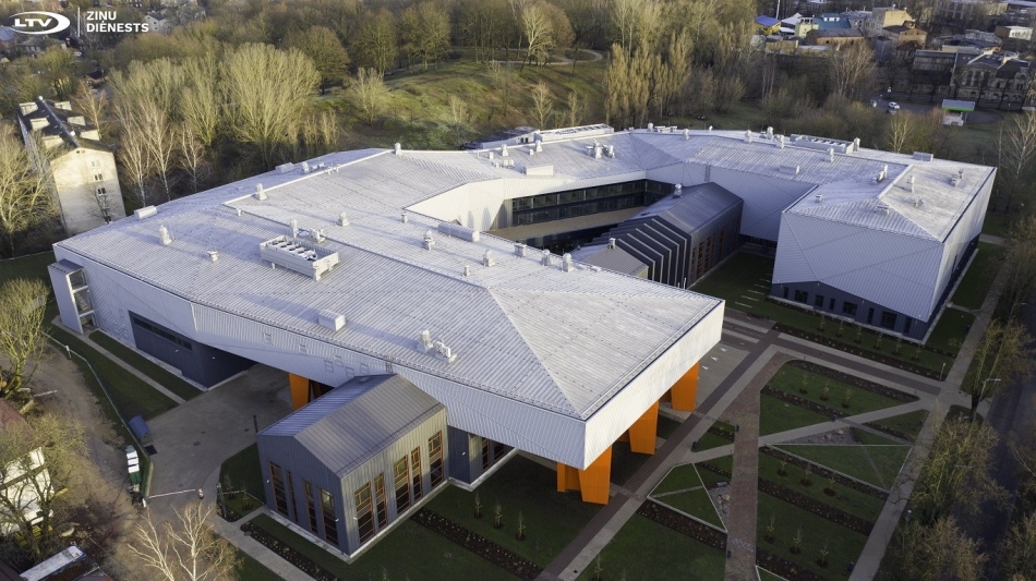 Luftaufnahme eines modernen Gebäudekomplexes mit markanter Dacharchitektur, umgeben von Grünflächen und Wegen.