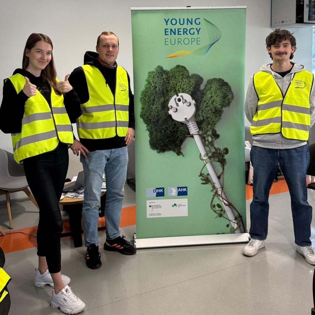 Drei Personen in gelben Warnwesten stehen neben einem Banner mit der Aufschrift ‚Young Energy Europe‘ und einem Bild eines Steckers im Grünen