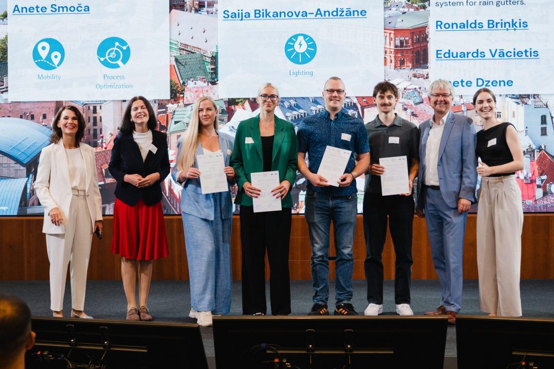 Acht Personen stehen auf einer Bühne und halten Zertifikate; im Hintergrund zeigt eine große Leinwand Namen und Themen wie ‚Mobilität‘ und ‚Prozessoptimierung‘