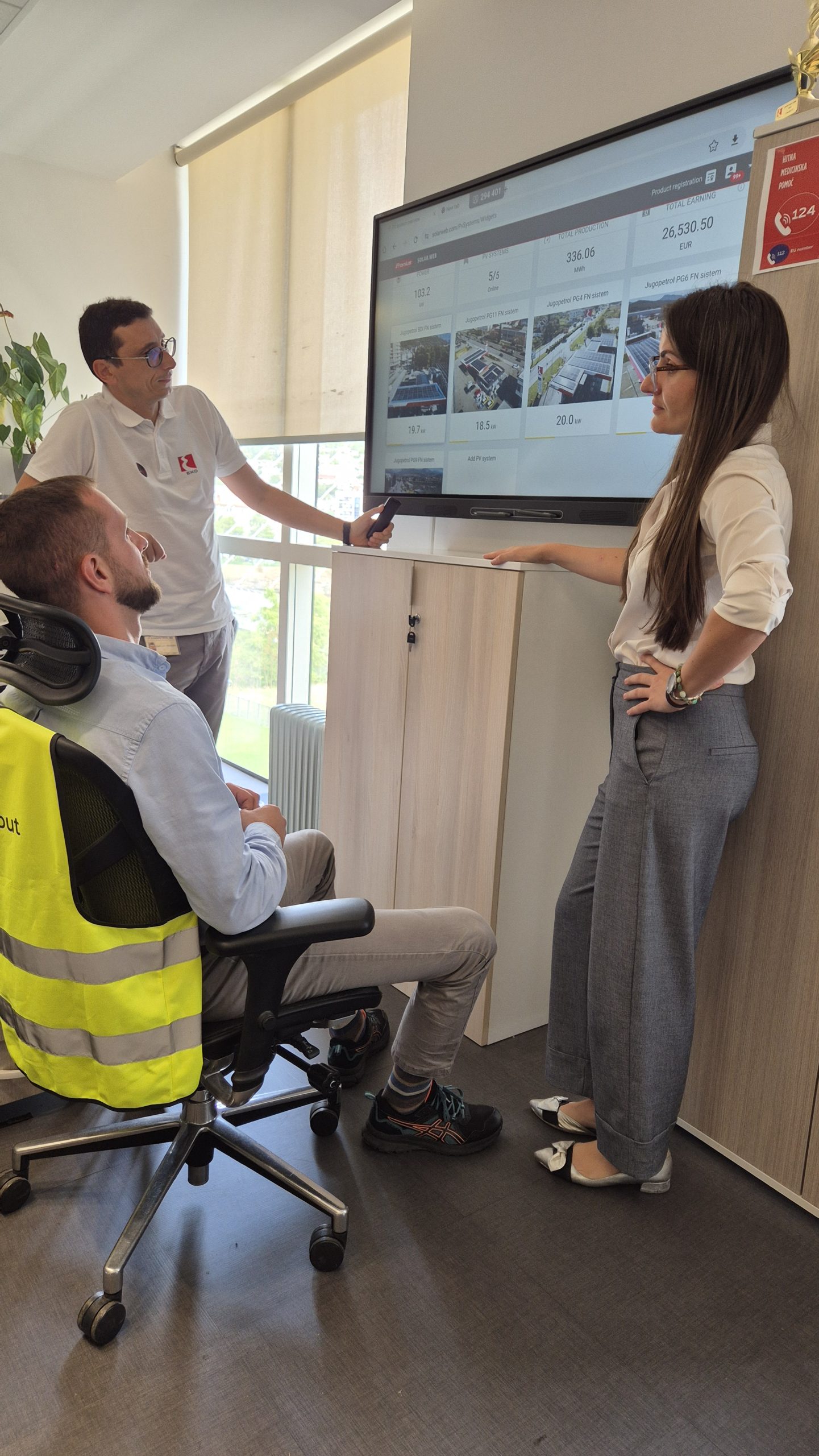 Three people (Energy Scouts) are in an office. One person is sitting, two are standing. They are looking together at a large screen on the wall. Numbers, images, and text can be seen on the screen. They are talking to each other.