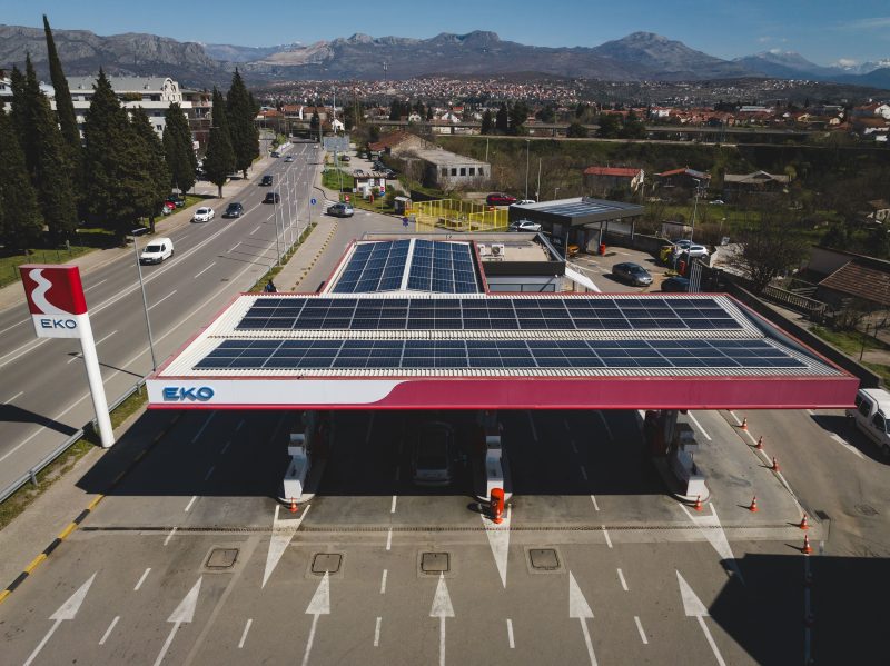 EKO gas station next to a multi-lane road. Solar panels are installed on the roof of the station. Mountains can be seen in the background behind the gas station.