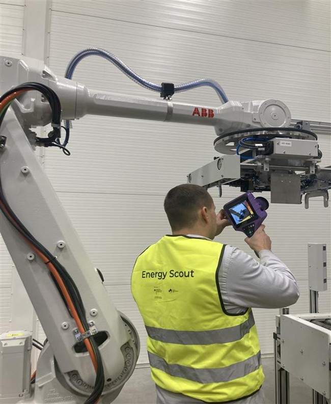 The image shows an Energy Scout wearing a yellow safety vest with the words “Energy Scout” printed on it, pointing a handheld measuring device at a large industrial robot. The robot arm is white and bears the ABB brand name, and the scene is set in a bright industrial hall with a white wall in the background. (©UAB HELLA Lithuania)