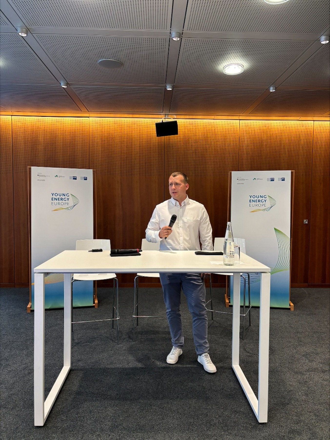 Das Bild zeigt einen Konferenzraum mit Holzvertäfelung und zwei Roll-up-Bannern mit dem Logo „Young Energy Europe“. Im Vordergrund steht ein weißer Stehtisch mit einer Wasserflasche, einem Glas, einem Tablet und einem kleinen Gerät; dahinter steht Energy Scout Paulius Majorovas. (©DIHK/Dirk Enters)