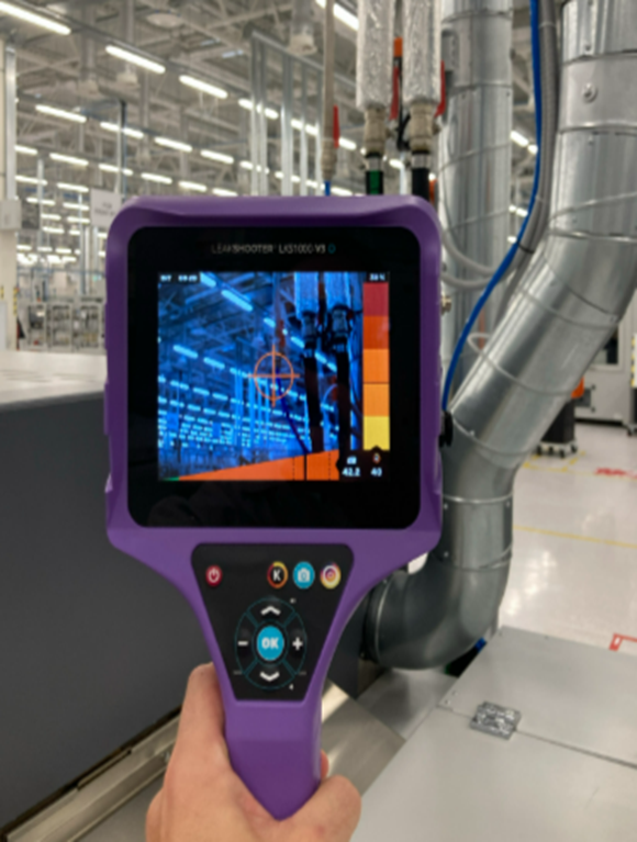 The image shows a handheld measuring device with a screen displaying a thermal image of an industrial hall. In the background are large metal pipes and parts of a technical system in a brightly lit production environment. (©UAB HELLA Lithuania)