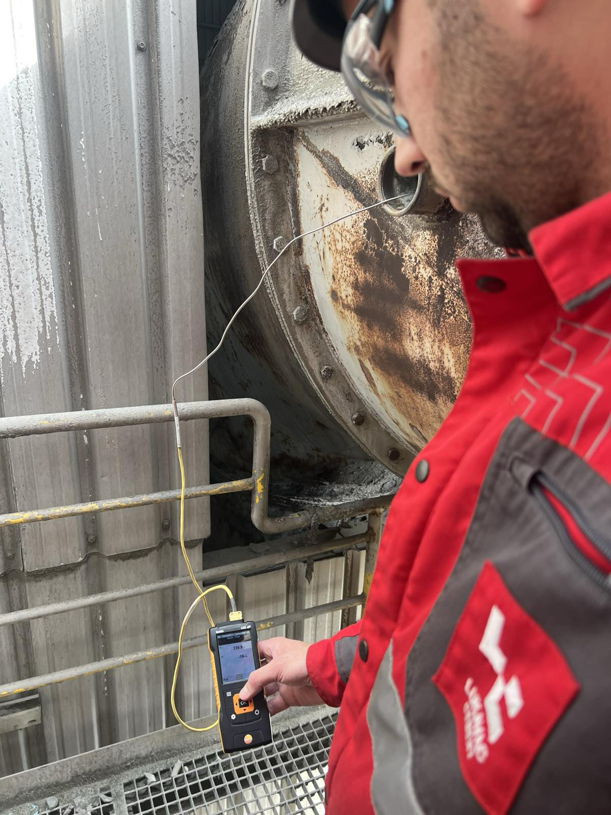 Person in roter Arbeitskleidung misst mit einem Handgerät und Kabel die Ablufttemperatur an einem großen, metallischen Industriebauteil, den Klinkerkühler.