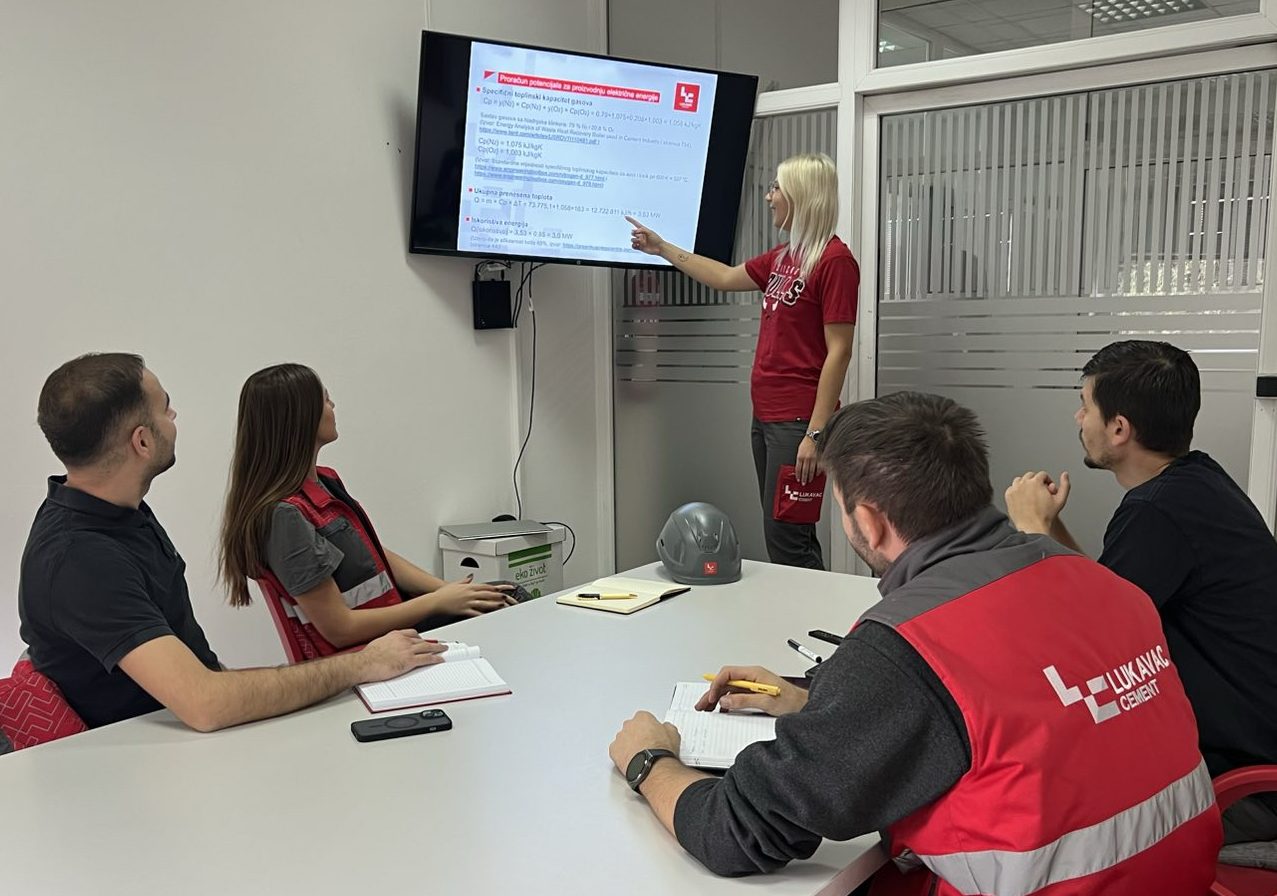 Person stands in front of a screen pointing at a presentation, while four other people sit at a table taking notes; notebooks, pens, and a safety helmet are on the table.