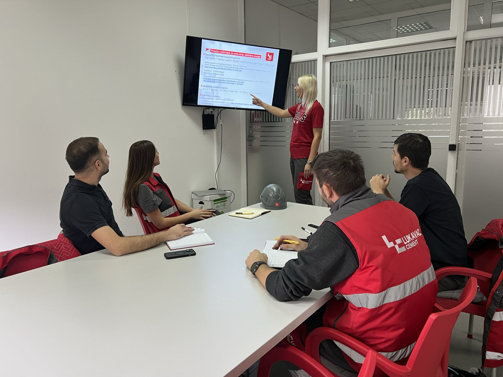 Eine Person steht vor einem Bildschirm und zeigt auf eine Präsentation, während vier weitere Personen an einem Tisch sitzen und Notizen machen; auf dem Tisch liegen Notizbücher, Stifte und ein Schutzhelm.