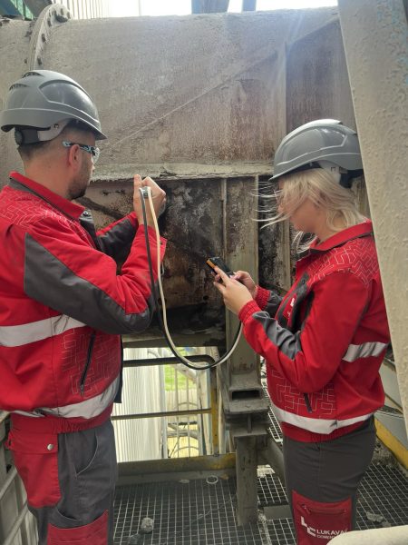 Two people in red workwear and safety helmets measure the airflow in the exhaust of the clinker cooler using devices with cables.