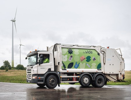 A white garbage truck with a green recycling-themed advertisement is parked on a wet road.