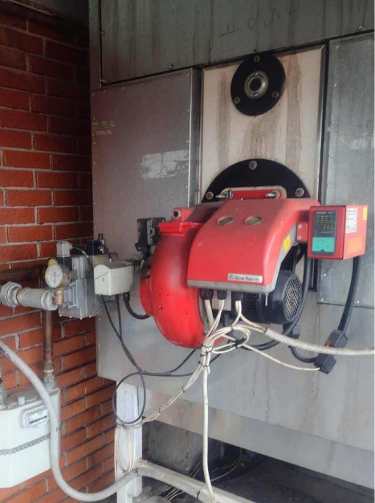 The image shows industrial heating equipment with a red burner unit mounted on a metal boiler surface, connected to several pipes, cables, and a control display.