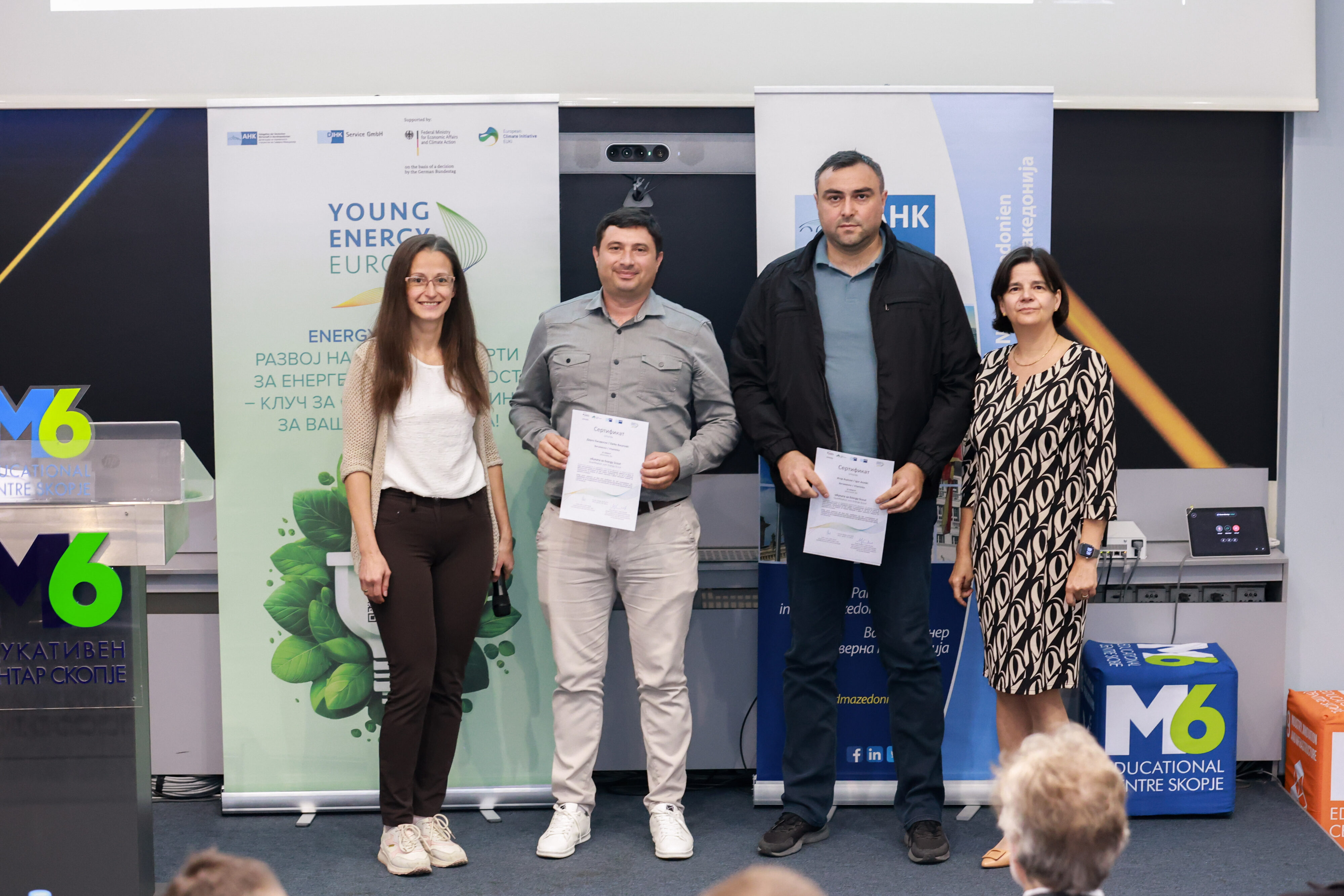 Four people are standing side by side indoors, with two of them holding certificates in front of project-related roll‑up banners. A lectern with the “M6” logo and a seating cube labeled “M6 Educational Centre Skopje” are visible in the room, along with an audience seated in the foreground.