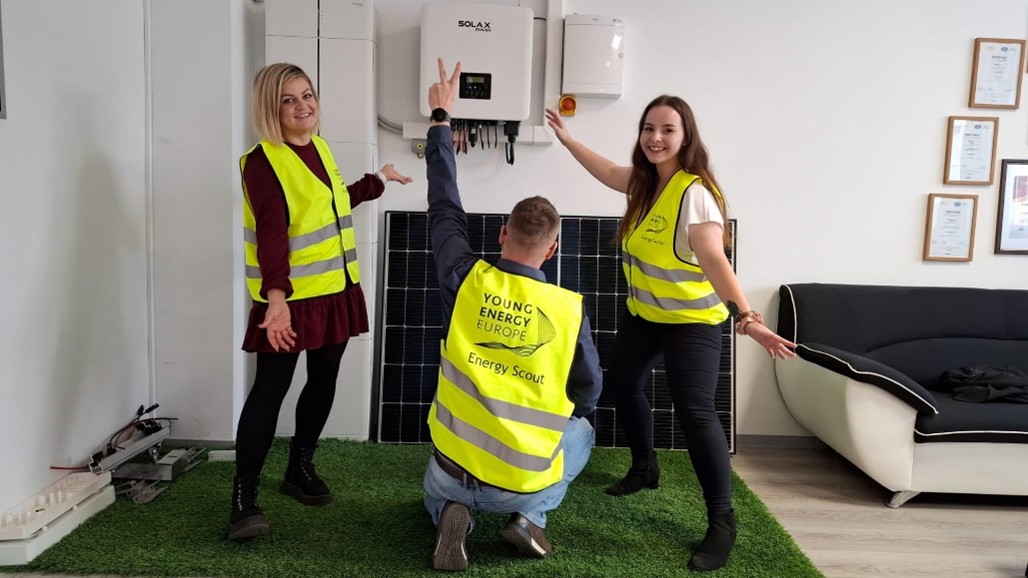 Three Energy Scouts in safety vests are standing indoors in front of a wall‑mounted inverter and photovoltaic panels, indicating an installed solar power system. The system is fully assembled and ready for operation, demonstrating the completed photovoltaic installation. (© Urbanix)