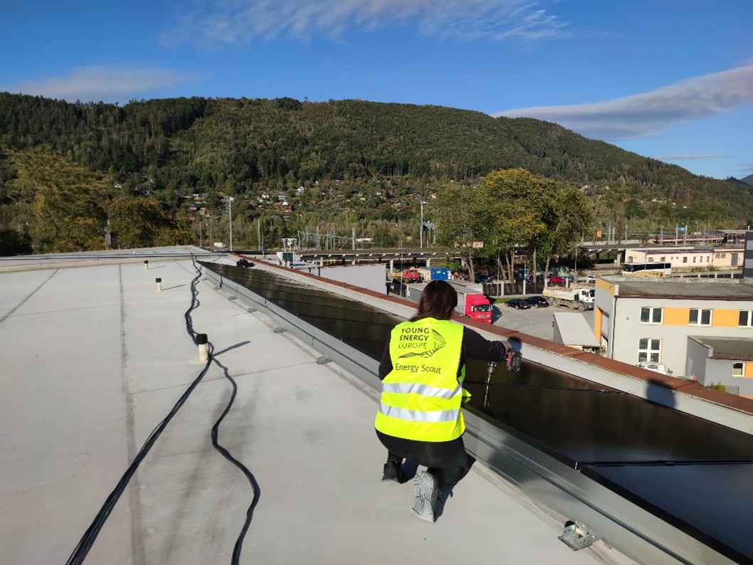 Das Bild zeigt eine Energy Scout in einer Warnweste, die auf einem Flachdach an der Installation einer Photovoltaikanlage mitarbeitet. Sichtbar sind Solarkabel, Montagematerial sowie umliegende Gebäude, die den praktischen Einsatz bei der Dachinstallation veranschaulichen. (© Urbanix)